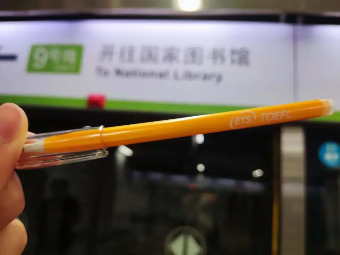 手持一支 ETS TOEFL 品牌橙色圆珠笔,背景为地铁开往国家图书馆的站台标识